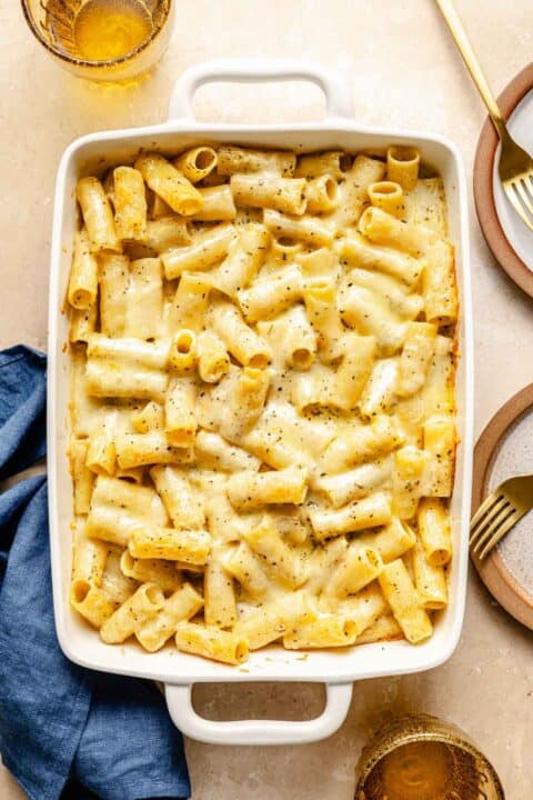 baked three cheese mac and cheese presented in a white baking pan next to small plates with gold forks and a blue linen towel