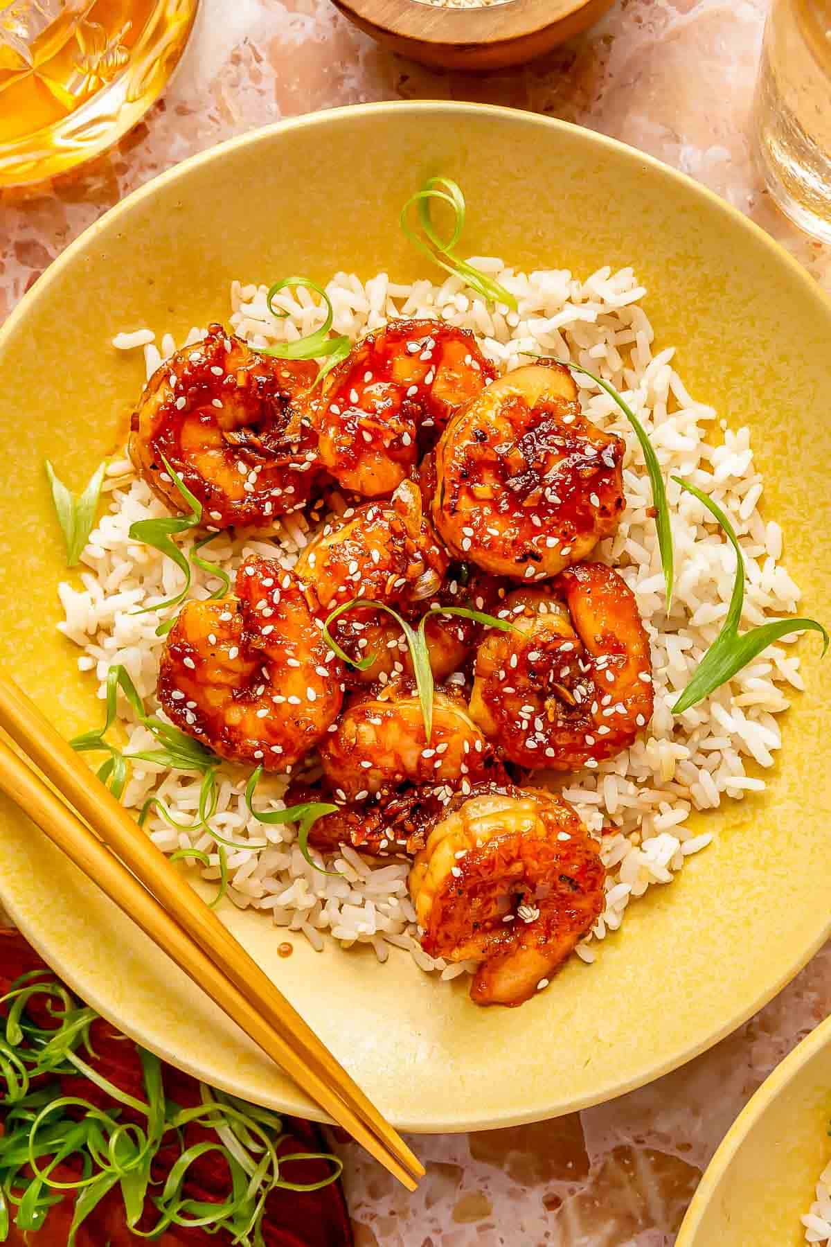 honey garlic shrimp on a bed of white rice in a yellow plate with wooden chopsticks and garnished with green onions and white sesame seeds.