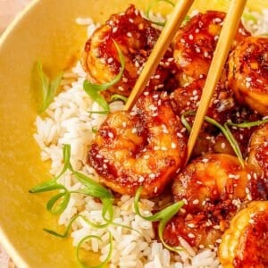 a pair of wooden chopsticks is holding a honey garlic shrimp that is on white rice