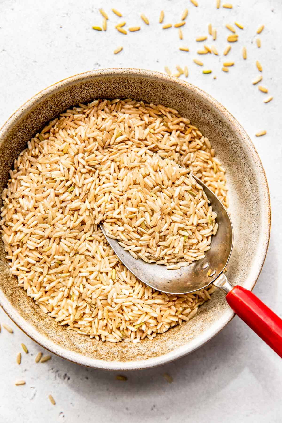 a metal scoop with a red handle is in the center of a brown bowl with uncooked brown rice grains