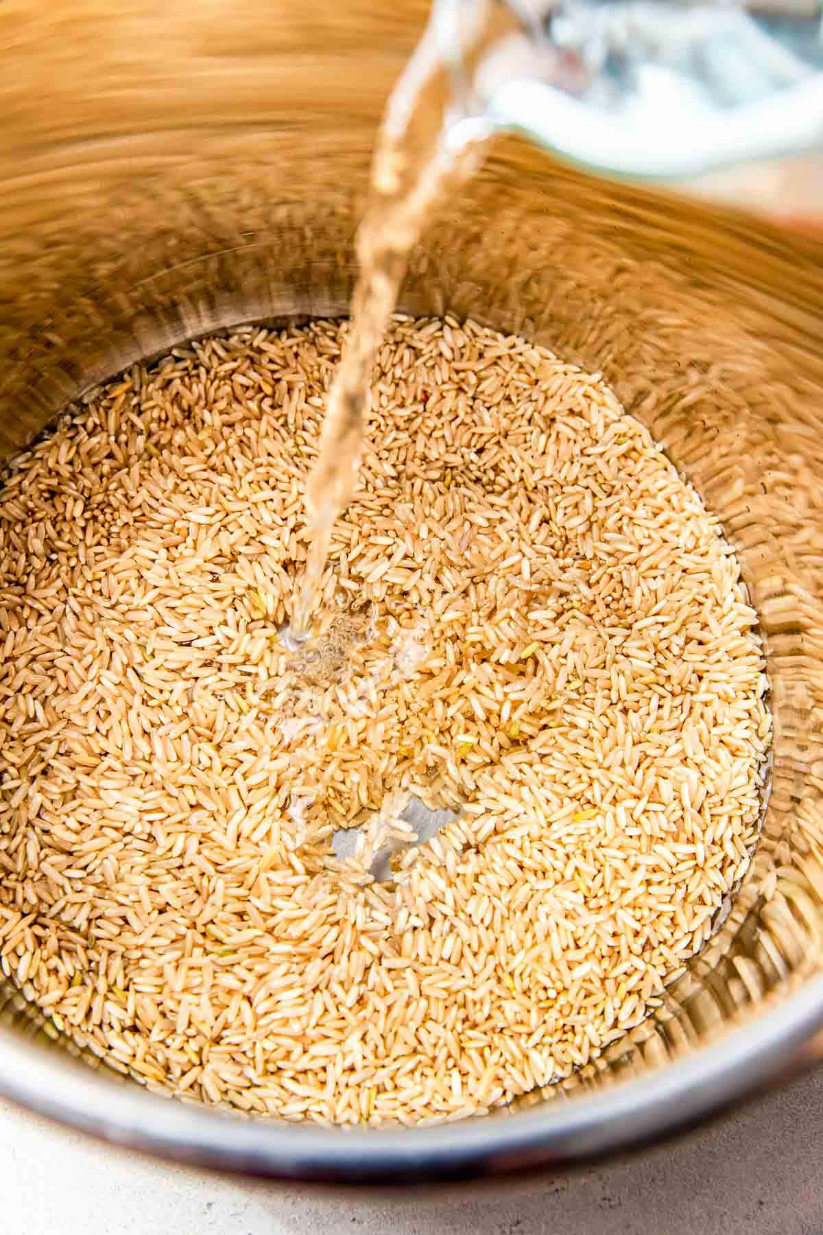 uncooked brown rice is inside a stainless steel pot with water being poured inside