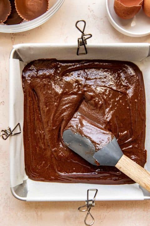 a grey silicone spatula is spreading brownie batter into a parchment paper lined square baking pan