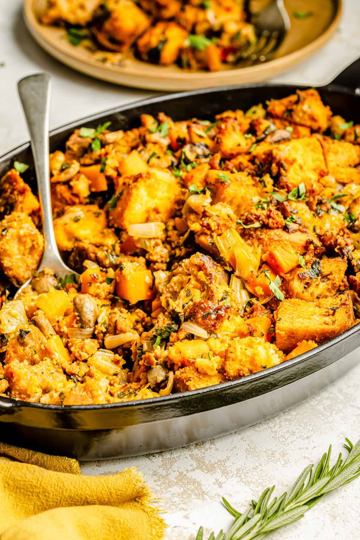 a metal spoon lies on the side of the chorizo cornbread stuffing in the oval baking dish next to fresh rosemary and a yellow tea towel