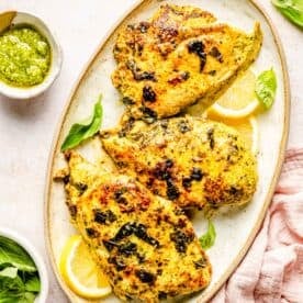 3 pieces of lemon basil chicken plated on a oval plate next to fresh pesto and a pink linen towel