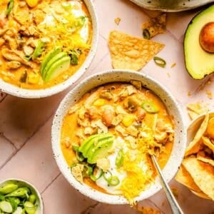 two bowls of southwest chicken chili topped with shredded cheese, avocado slices, sour cream, and tortilla chips