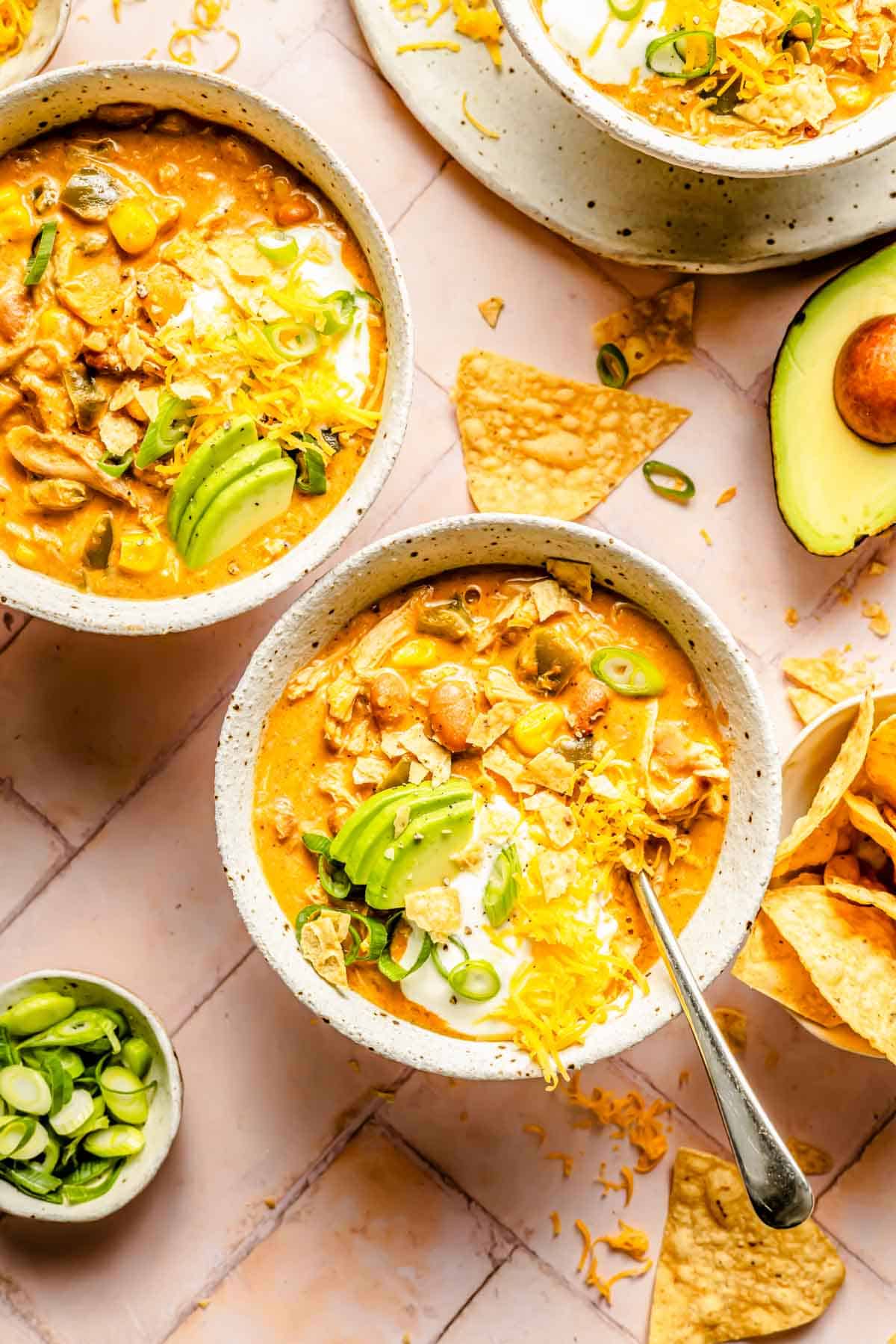 two bowls of southwest chicken chili topped with shredded cheese, avocado slices, sour cream, and tortilla chips