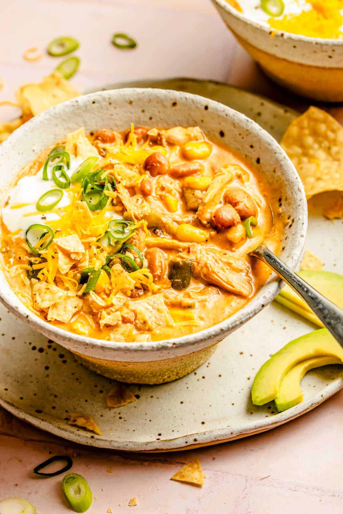 a bowl of southwest chicken chili is in a bowl with a metal spoon and on a plate with sliced avocados