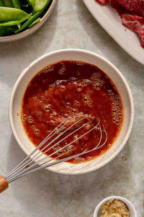 A bowl filled with a reddish-brown marinade with visible bubbles on the surface. A whisk is placed in the bowl. Nearby are small bowls with chopped garlic and a plate with green vegetables and sliced raw meat.