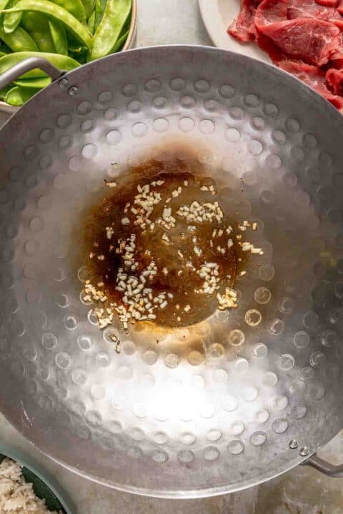 A large gray wok on a stovetop contains oil and minced garlic being sautéed. Surrounding the wok are plates with green vegetables, raw sliced beef, and a bowl of rice.