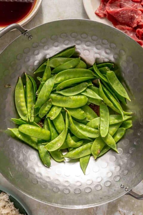 A large metal wok containing fresh snap peas sits on a countertop. Nearby, there are bowls with ingredients, including sliced raw meat and a dark liquid sauce. A plate of rice is partially visible in the corner.