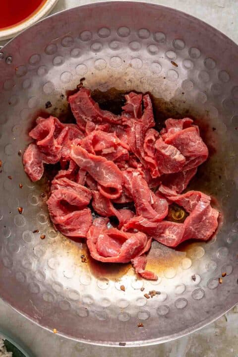 Slices of raw beef are placed in a large metal wok, ready for cooking. The wok has a textured surface, and some soy sauce is visible at the bottom.
