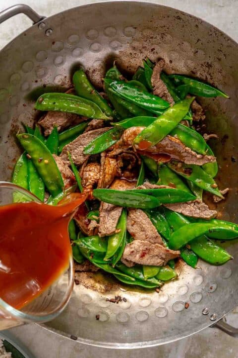 A wok containing a stir-fry of sliced beef and snap peas. A sauce is being poured over the ingredients from a glass measuring cup.