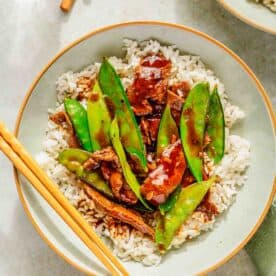 A bowl of rice topped with stir-fried beef, green snow peas, and a savory sauce. Chopsticks are placed beside the bowl on a light-colored surface. A glass of water is partially visible in the background.