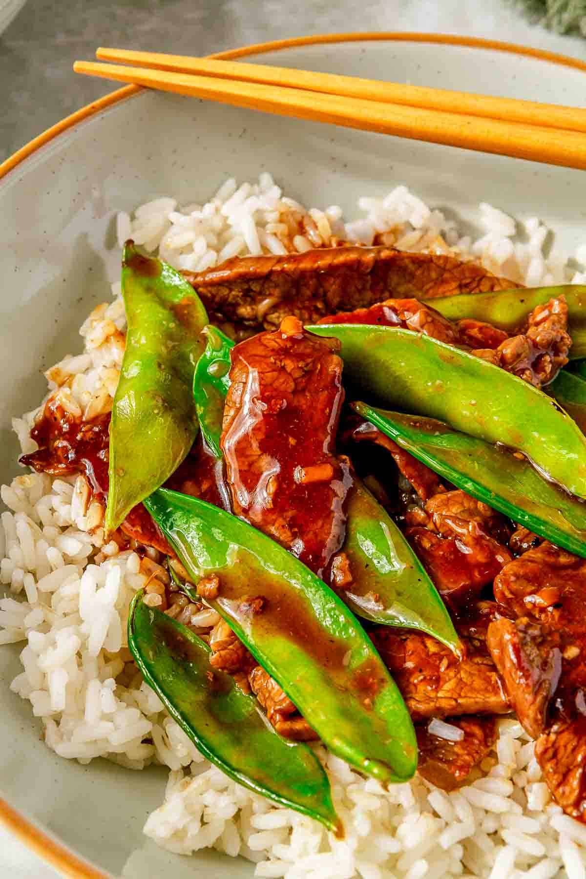 A bowl of white rice topped with slices of beef and green snow peas, coated in a dark sauce. Chopsticks are placed on the rim of the bowl.