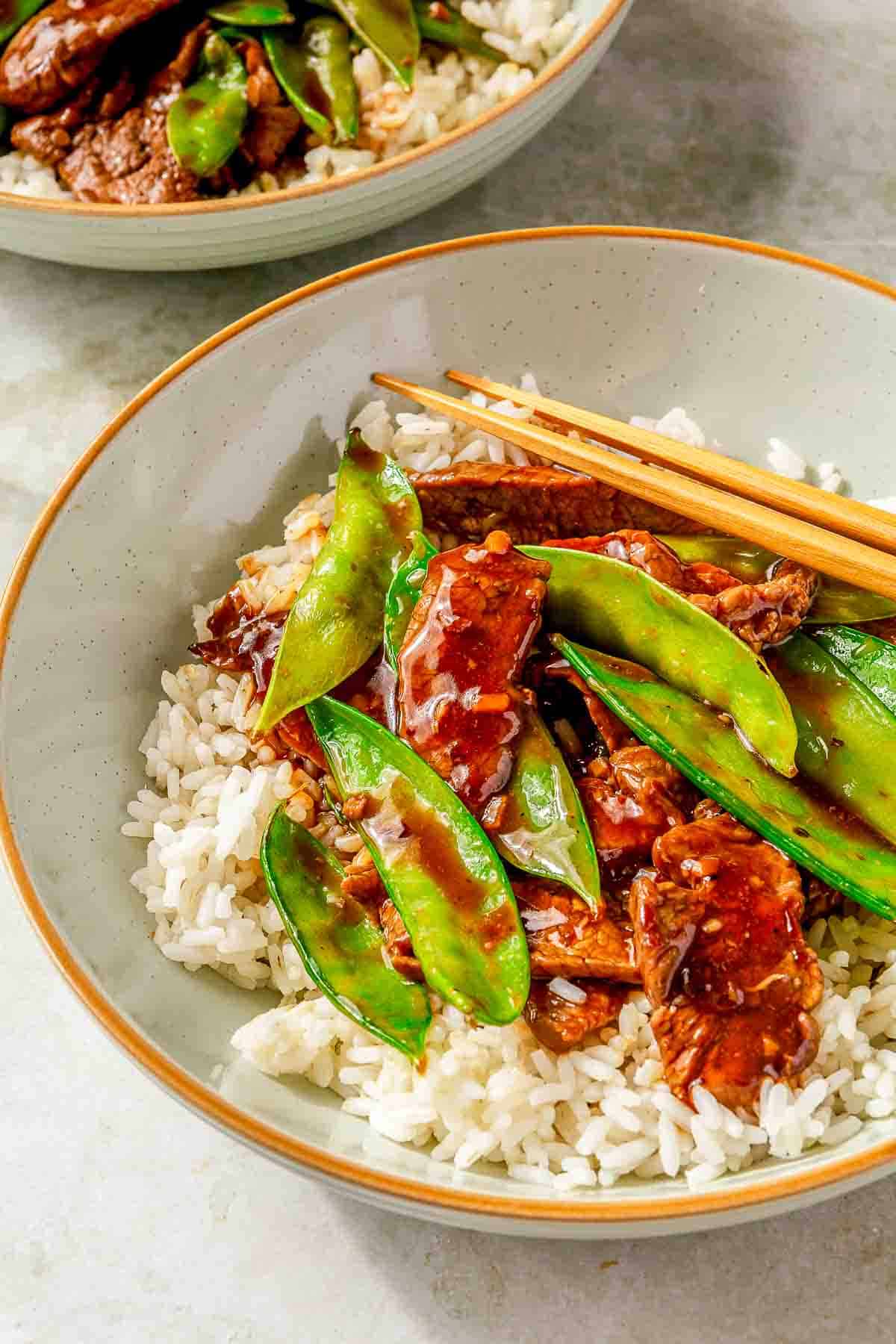 A bowl of beef stir-fry with snow peas and sauce on a bed of white rice. Chopsticks rest on the rim of the bowl. Another bowl of a similar dish is partially visible in the background.