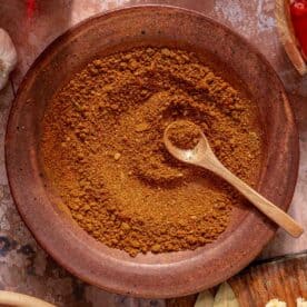A brown ceramic bowl filled with a reddish-brown spice blend. A wooden spoon rests inside the bowl. Surrounding items include raw meat, garlic cloves, bay leaves, and chopped onions on a textured surface.