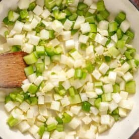 Close-up of a pot with diced onions, celery, and green bell peppers being stirred with a wooden spoon. The vegetables are uncooked, showing a mix of white and green colors. Carrot slices can be seen at the edge of the image.