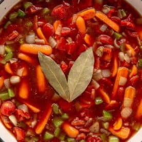 A close-up of a pot filled with colorful vegetable soup, featuring chopped tomatoes, carrots, celery, onions, and two bay leaves on top. The broth is rich and red, with various vegetables visibly submerged.