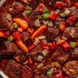 A close-up of a pot filled with beer braised pot roast. The stew contains chunks of beef, carrots, celery, onions, and diced tomatoes in a rich, brown broth. The colorful ingredients are mixed together, creating a hearty and appetizing appearance.