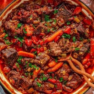A savory beer braised pot roast in a green pot, filled with chunks of beef, carrots, tomatoes, and herbs. A wooden spoon rests on the side. Fresh parsley is scattered on top, and a glass of red wine and a bunch of parsley are beside the pot.