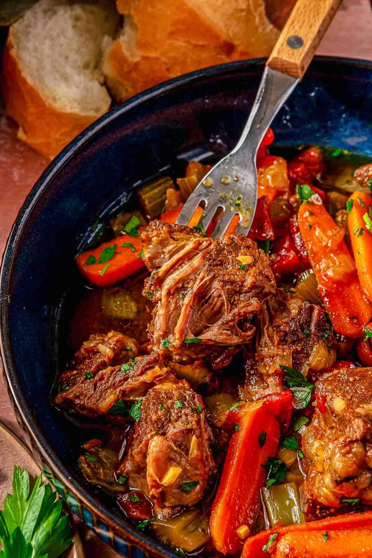 A close-up of beer braised pot roast in a blue bowl, featuring tender chunks of beef and vibrant carrots. The dish is garnished with fresh herbs and accompanied by slices of bread in the background, creating a warm and hearty presentation.