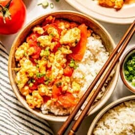 Bowl of rice topped with scrambled eggs and tomatoes, garnished with green onions, next to whole tomatoes and a striped napkin. Chopsticks rest on the bowl.