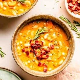 A bowl of bean soup topped with diced bacon and garnished with rosemary, next to a bowl and spoon.