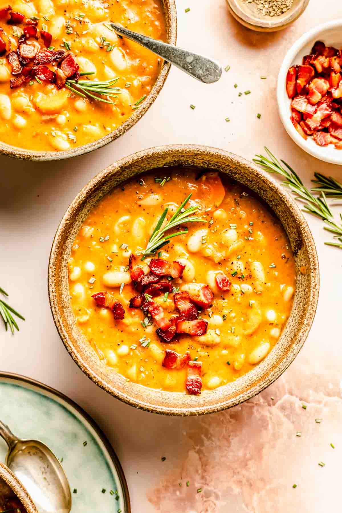 A bowl of bean soup topped with diced bacon and garnished with rosemary, next to a bowl and spoon.