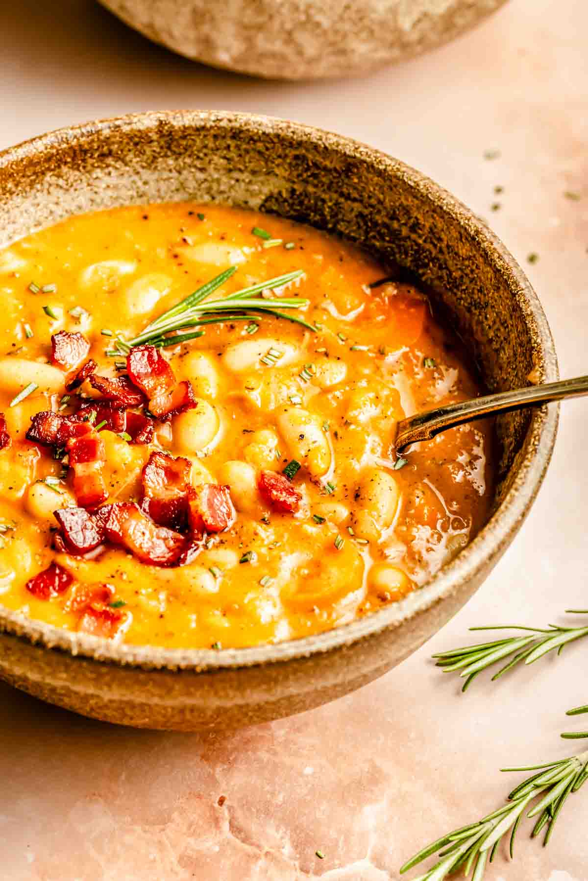 A bowl of white bean soup topped with diced bacon and rosemary, with a spoon on the right.