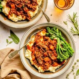 Two bowls of mashed potatoes topped with short rib ragu, garnished with green herbs and broccolini, next to a beige napkin and glasses of amber liquid.