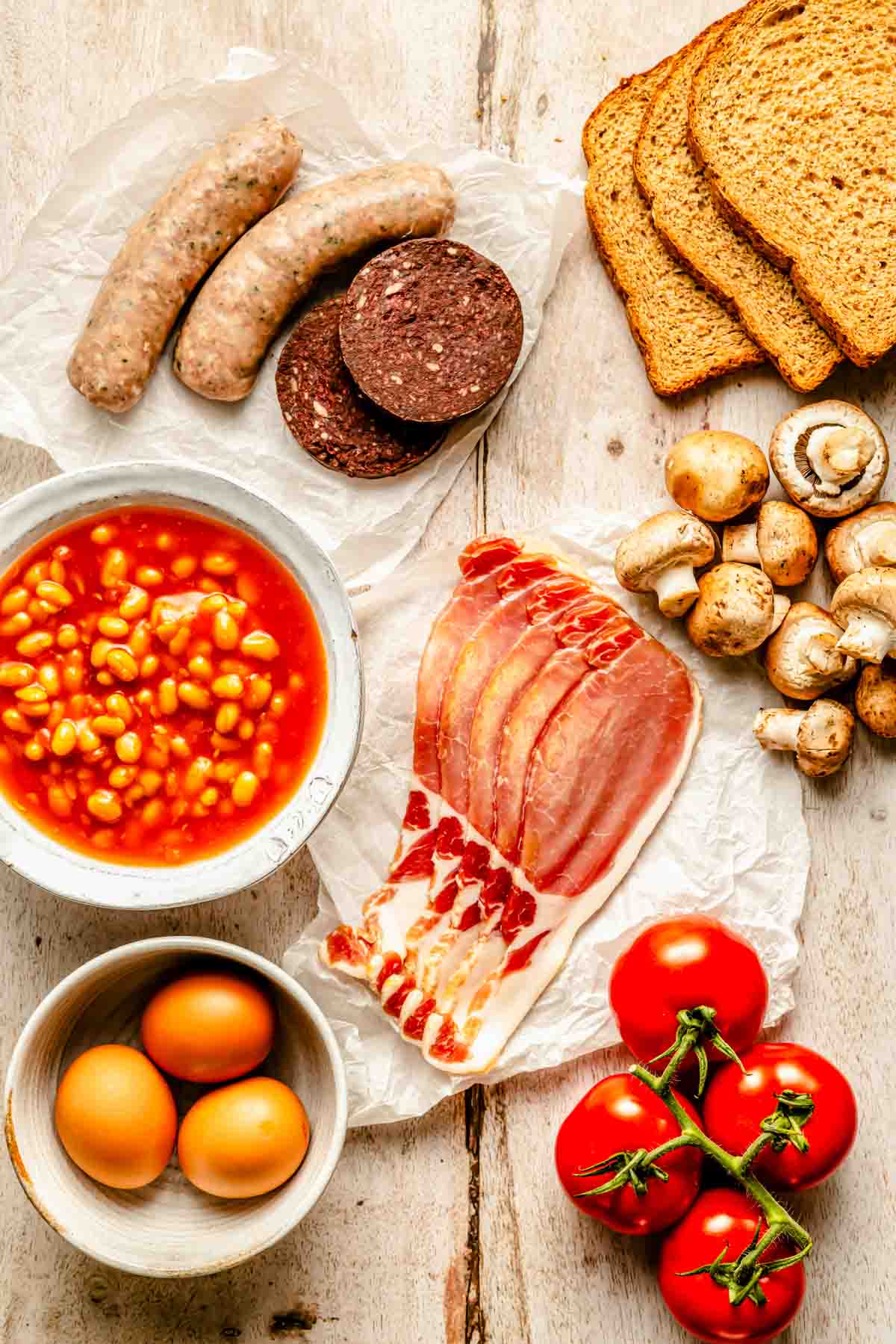 Ingredients for a full English breakfast: sausages, black pudding, baked beans, back bacon, eggs, tomatoes, mushrooms, and sliced bread laid out on a wooden surface.