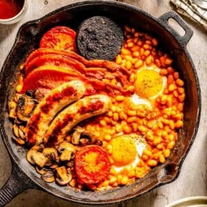 A cast iron skillet with baked beans, fried eggs, sausages, back bacon, black pudding, mushrooms, and grilled tomatoes on a wooden table.