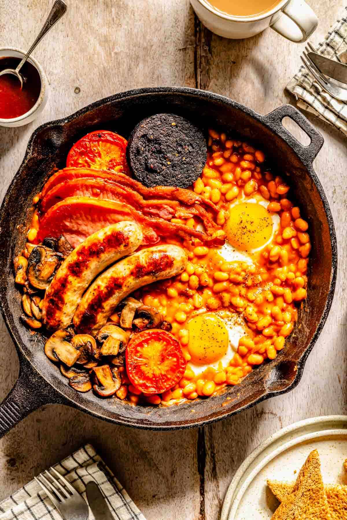 A cast iron skillet with baked beans, fried eggs, sausages, back bacon, black pudding, mushrooms, and grilled tomatoes on a wooden table.