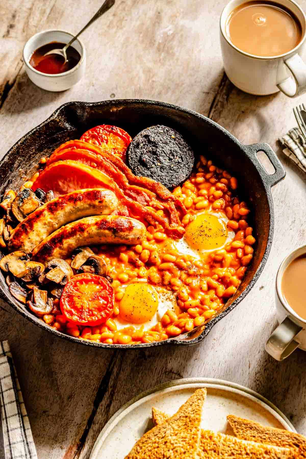 A skillet with eggs, beans, sausages, back bacon, tomatoes, mushrooms, and black pudding. Toasted bread and cups of tea on the side.