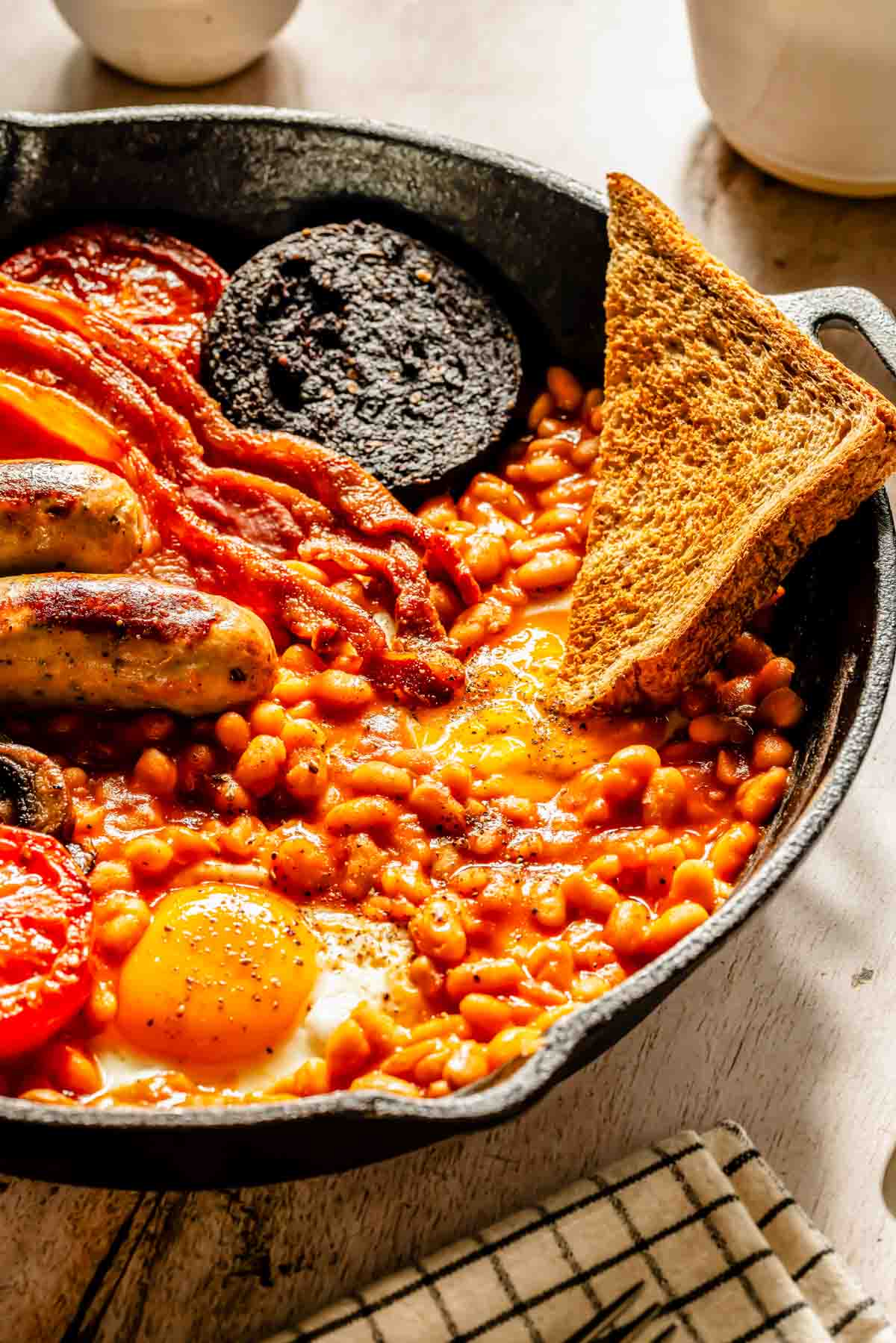 A cast iron skillet with baked beans, eggs, sausages, back bacon, black pudding, tomato, and a slice of toast on a wooden table.