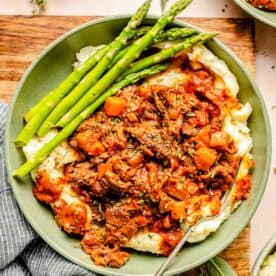 A plate of mashed potatoes topped with lamb ragu and served with asparagus. A napkin and herbs are on the table beside the dish.