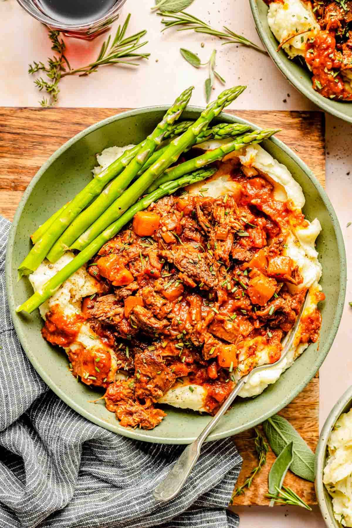 A plate of mashed potatoes topped with lamb ragu and served with asparagus. A napkin and herbs are on the table beside the dish.
