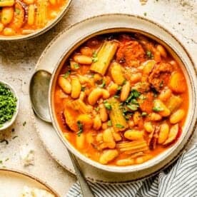 A bowl of soup with beans, sausage, and celery, garnished with herbs. A spoon rests beside the bowl, and pieces of bread are scattered around on a striped linen towel.