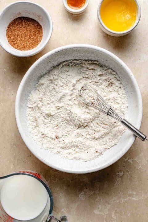 A bowl of flour with a whisk inside is surrounded by small bowls and a measuring cup containing cinnamon, vanilla, melted butter, and milk on a beige countertop.