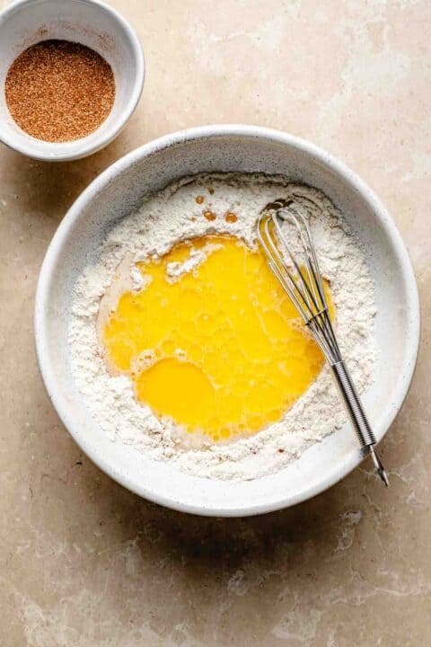 Bowl of flour with melted butter and a whisk. A small bowl of cinnamon sugar is next to it on a beige surface.