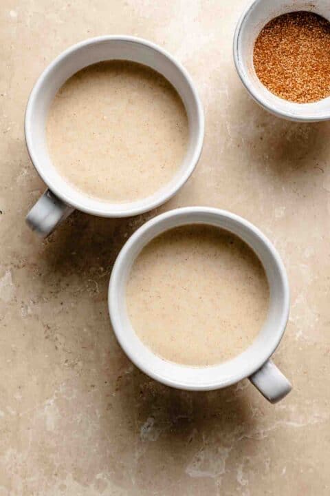 Two white mugs filled with batter on a marble surface, accompanied by a small bowl of cinnamon sugar nearby.