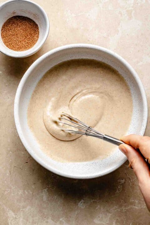 Person whisking batter in a bowl next to a small bowl of cinnamon sugar on a marble surface.