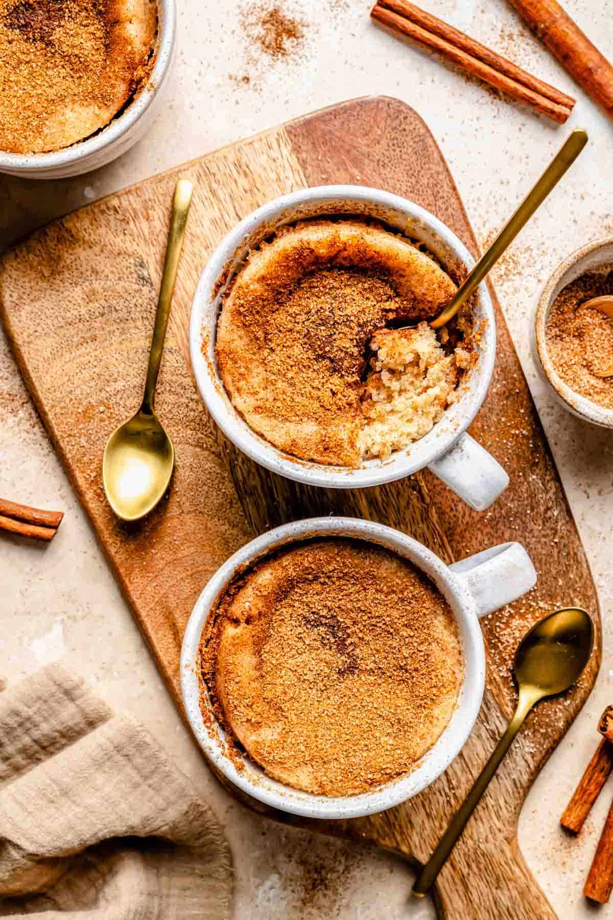 Two mugs of snickerdoodle mug cake on a wooden board with spoons and cinnamon sticks nearby.