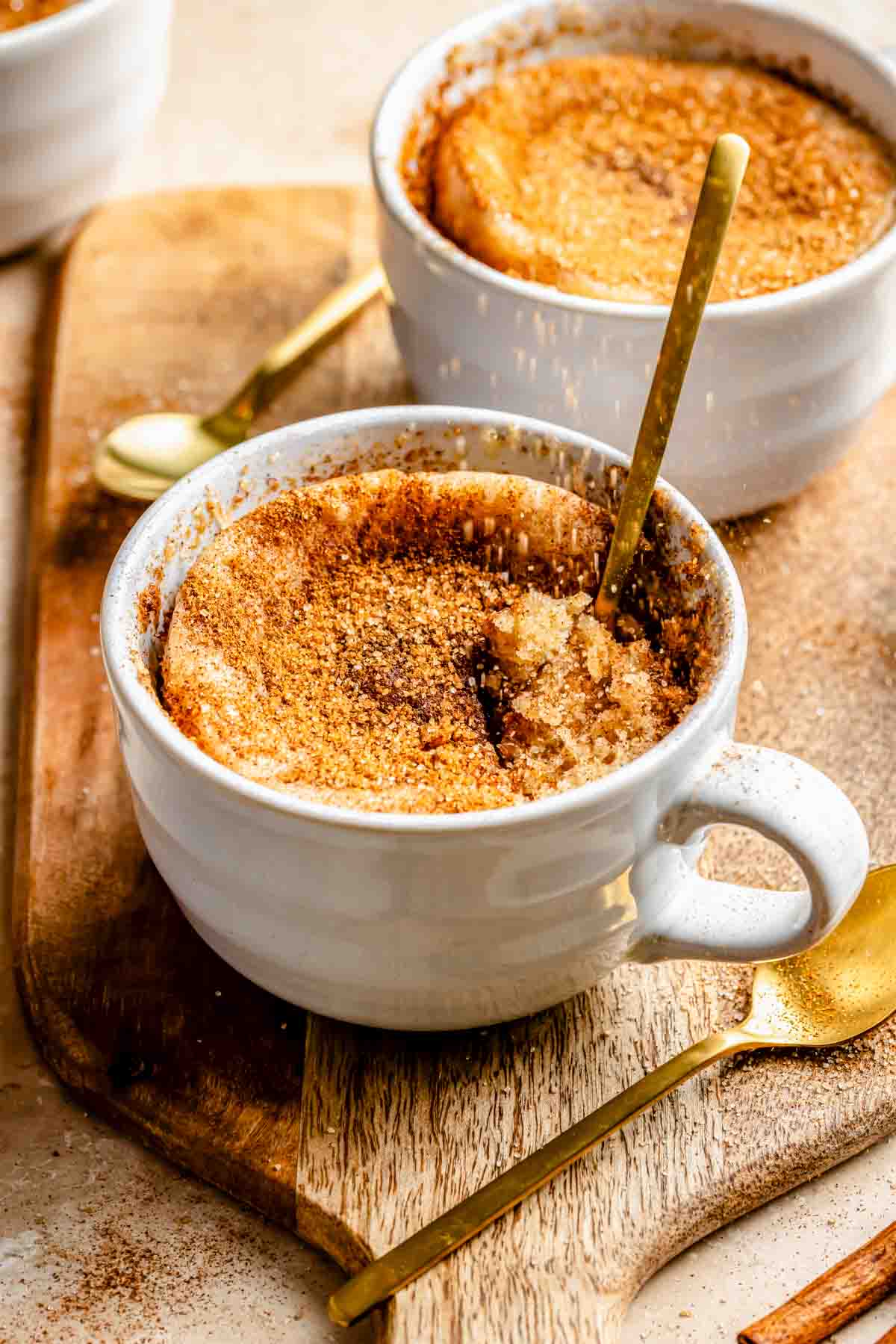 Two white ceramic cups filled with snickerdoodle mug cake, cinnamon sugar-topped, with spoons. The cups are placed on a wooden board with an additional gold spoon beside them.