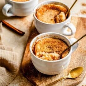 Three white mugs filled with snickerdoodle mug cakes on a wooden board, accompanied by gold spoons and cinnamon sticks.