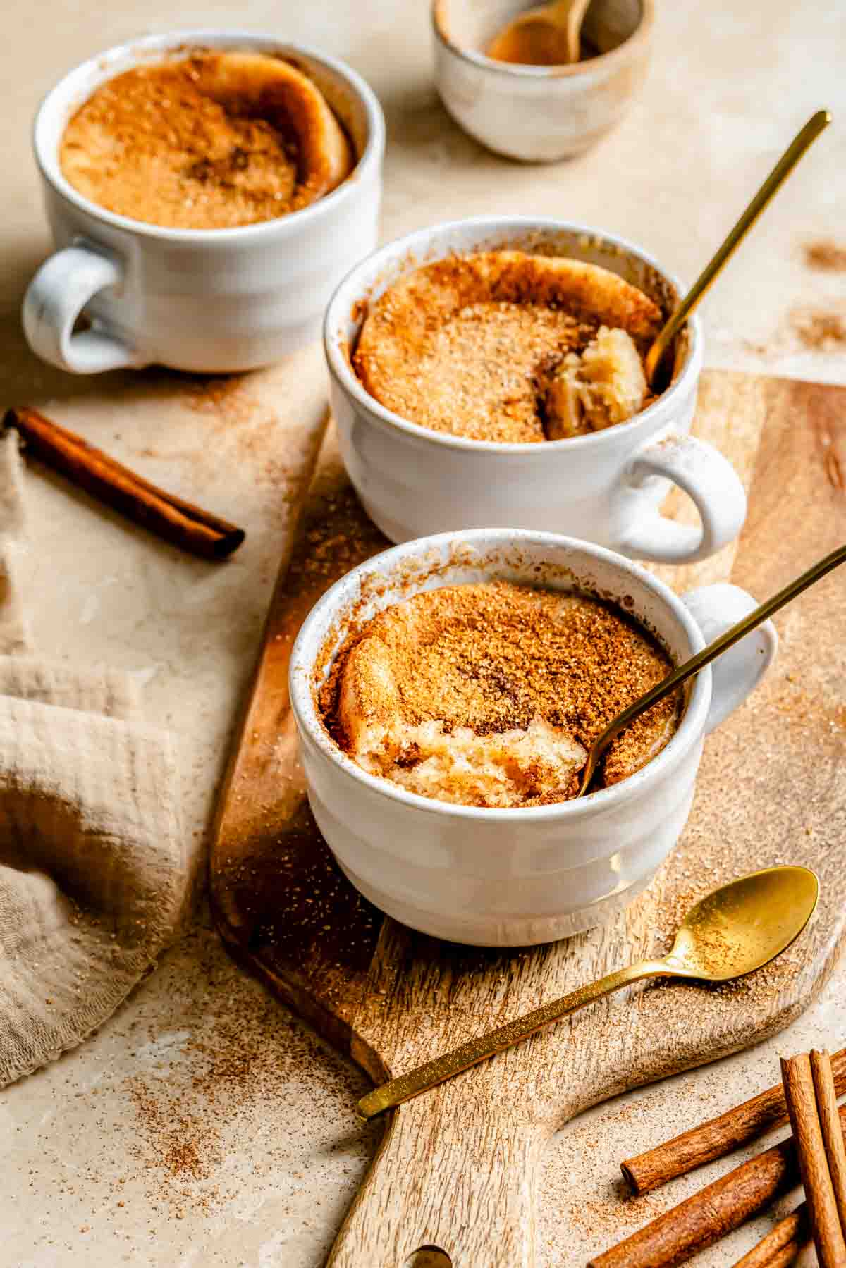 Three white mugs filled with snickerdoodle mug cakes on a wooden board, accompanied by gold spoons and cinnamon sticks.