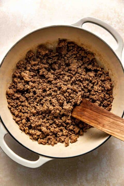 A pot filled with cooked, crumbled ground beef and sausage, with a wooden spatula resting inside.