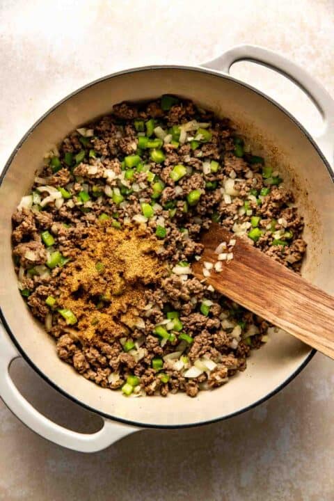 A pot with cooked ground meat, chopped green peppers, onions, and spices, with a wooden spatula.