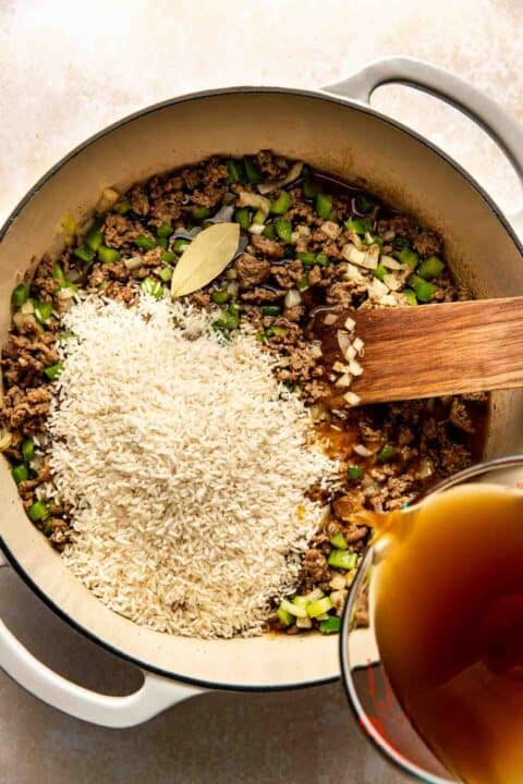 A pot containing uncooked rice, ground meat, chopped green peppers, onions, and a bay leaf is being cooked. A wooden spoon stirs the mixture, while broth is being poured in.