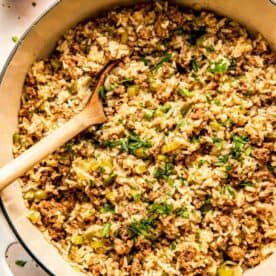 A pot of dirty rice mixed with ground meat, green bell peppers, and herbs, with a wooden spoon resting inside.